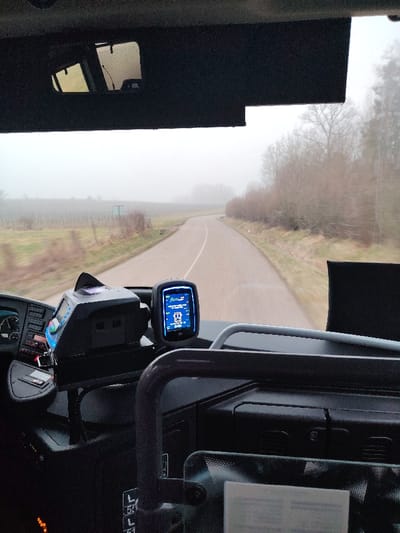 A view from inside a vehicle, likely a bus or truck, shows a paved road winding through a foggy landscape. Bare trees and sparse vegetation line the roadside. The dashboard features electronic devices, including a GPS unit displaying information. A rearview mirror is visible. The overcast conditions create a muted color palette with the road and sky blending into a hazy backdrop.