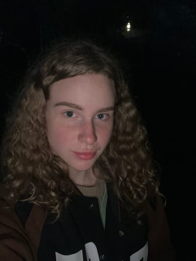 A portrait of a young individual with light skin, blue eyes, and curly brown hair. They are wearing a dark jacket with a light-colored letter on the chest over a lighter-colored sweater. The background is mostly dark, with a few indistinct lights in the upper left corner. The location is possibly Krasnyi Jar, Russia, as indicated in the query, though not visually verifiable.
