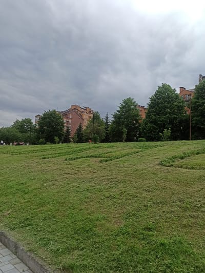 The image depicts a slightly overcast day in what appears to be a city park or residential area, with a large, freshly mown grassy slope in the foreground. The slope is covered in a thick layer of cut grass, indicating recent maintenance. A paved walkway runs along the bottom left edge of the image, bordered by a concrete curb.
In the middle ground, behind the slope, a line of mature trees with lush green foliage is visible. Beyond the trees, a multi-story brick building with a light orange facade and multiple windows is situated. Some of the windows appear to have air conditioning units. The building's architecture suggests it is an apartment complex or office building. Several other trees are scattered around the building, and further to the right, more of the same type of building is partially visible.
In the far background, the sky is filled with heavy, gray clouds, suggesting potential rain or just a generally cloudy atmosphere.
On the left side of the image, near the trees and in front of the building, a few people are casually walking and sitting on benches, engaged in light activity. One person is wearing an orange vest, possibly a park worker. The overall impression is a peaceful, urban environment undergoing seasonal upkeep. The location is stated to be in Atabe, Georgia, though this specific context is not visually verifiable from the image alone.