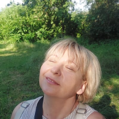 This media file features a close-up, overhead shot of a woman with blonde hair and closed eyes, lying on a grassy area in what appears to be a park or natural setting. The woman is smiling with her mouth slightly open, revealing small, sparkly dental embellishments on her teeth, likely rhinestones or similar decorative items. She is wearing a striped top with a light-colored strap that has a metal buckle. The background consists of lush green grass and trees, suggesting an outdoor environment during daylight. The lighting indicates it could be late afternoon due to the warm, golden tones reflecting on the woman's face. There are no visible text elements in the image. The overall impression is one of relaxation and contentment in a natural, sunlit environment, consistent with a location context of Voronkiv, Ukraine.