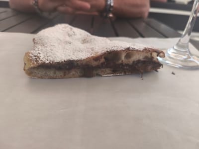 A close-up, slightly overhead shot shows a slice of dessert, possibly a calzone or pastry, filled with chocolate and dusted with powdered sugar. The dessert rests on a sheet of white paper. In the background, blurred figures of people are visible, along with a table and a clear glass. The scene appears to be outdoors, possibly at a cafe or restaurant in San Salvo, Italy. The lighting is soft and natural.