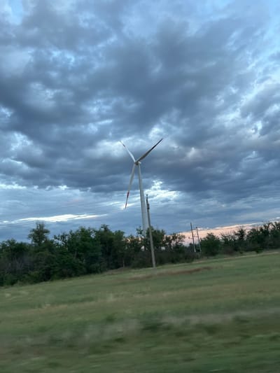 An outdoor, eye-level, medium shot captures a wind turbine under a dramatic, overcast sky, a classic view of Zimovniki, Russia. The turbine, white with red accents at the tips of its blades, stands tall in the center of the frame. Its three blades are partially obscured by the cloudy sky. A field of green grass, slightly blurred by motion, fills the foreground. In the middle ground, a line of dense green trees runs horizontally across the image, with power lines and poles visible above them. The sky is dominated by dark, textured clouds, suggesting a potentially stormy or twilight atmosphere. The overall impression is one of vast, open landscape and a powerful, modern energy source against a natural backdrop.