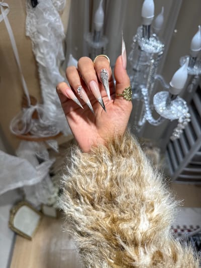 A hand adorned with long, stiletto-shaped nails featuring a nude base with silver and white intricate designs, including rhinestones. The hand wears a large gold ring on the index finger and a simpler gold band on the pinky. The hand is positioned in front of a furry, beige garment. A chandelier is visible in the blurred background, alongside some white fabric and a small, decorative object.