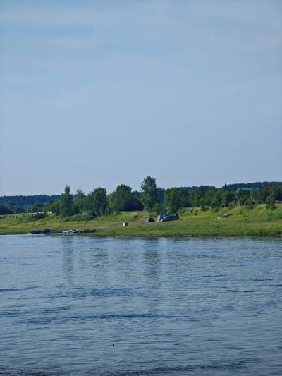 This is an image of a riverbank in Galanino, Russia. The foreground is dominated by a body of blue water with ripples. The middle ground shows a grassy riverbank lined with green trees. Several boats are pulled up onto the bank, some partially submerged. There are also a few tents and what appears to be a car parked on the grass. In the background, a dense line of trees stretches across the horizon, with a few small buildings visible amongst them. The sky is a clear, pale blue.