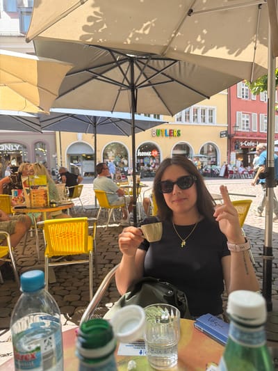 This image depicts an outdoor cafe scene on a sunny day in Freiburg im Breisgau, Germany. In the foreground, a woman wearing sunglasses and a black t-shirt is seated at a table, holding a small cup of coffee in her left hand. Her right hand is raised, with her index finger and thumb touching. She is wearing a gold pendant necklace and a pink watch.
The table is set with a glass of water, a blue pack of Vogue cigarettes, and partially visible bottles of water. The background shows other cafe patrons seated at yellow chairs under umbrellas. Beyond the cafe, buildings with yellow and red facades are visible, some featuring arched windows. The ground is paved with cobblestones. Signs in German, such as "BUTLERS" and "home24", are visible on the buildings, suggesting this is a commercial area within the city. The overall impression is of a relaxed, urban outdoor dining experience.