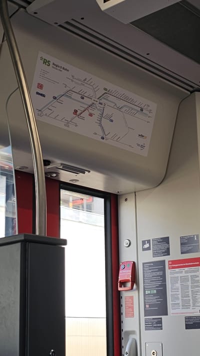 The image shows the interior of a train car, likely in Neu-Ulm, Germany, given the presence of German text on the signage. In the upper portion of the image, a map of the "RS Regio S-Bahn Donau-Iller" is displayed, indicating train routes and stations. Below the map, a red emergency brake handle is visible on a white panel, labeled "NOTBREMSE" in red text. To the right of the emergency brake, there are several informational posters. One poster, with the title "Ihre Fahrgastrechte im Eisenbahnverkehr," provides information about passenger rights in rail transport. The overall environment appears to be a functional, public transportation setting.