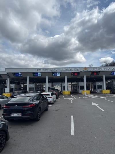 The image displays a multi-lane vehicle border crossing at Kalotina, Bulgaria, under an overcast sky. A line of cars is queued, facing forward towards the inspection booths. In the immediate foreground, the rear of a dark-colored BMW X4 M40i, bearing Georgian license plate GE-137852, is visible, positioned in one of the vehicle lanes. Behind it, a white sedan and other vehicles extend in a queue.

Overhead, a large, grey canopy spans the width of the crossing, supported by multiple pillars. The canopy's facade features "РЕПУБЛИКА БЪЛГАРИЯ" and "REPUBLIC OF BULGARIA" in prominent lettering, accompanied by the Bulgarian coat of arms. Below this main sign, above each lane, individual electronic display signs indicate lane status and vehicle type, reading "ALL PASSPORTS" and "CARS." Some lanes show green arrows, indicating they are open, while others display red 'X' symbols, indicating closure.

The asphalt road surface features clear white painted markings, including directional arrows and the word "CAR" designating the lanes. Yellow and black striped concrete barriers separate the individual inspection lanes. No specific individuals are clearly visible outside of being occupants within the vehicles. In the background, beyond the border infrastructure, a distant highway and sparse trees are discernible under the cloudy conditions.