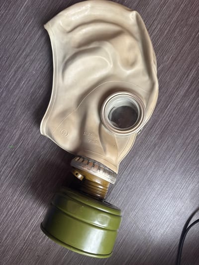 A gas mask is lying on a dark wooden surface. The gas mask is beige and has a green filter attached to the bottom. There are some markings on the gas mask, but they are not clear enough to read. The surface appears to be a tabletop or desk. There is no information in the image that indicates a location in the city of Otradny, Russia.