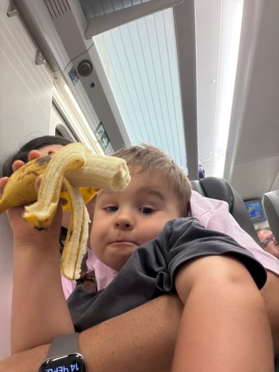 In this image, a young child with light brown hair is looking at the camera while holding a partially peeled banana. The child is wearing a pink shirt and a grey t-shirt. A person's arm, also wearing a grey t-shirt, is visible in the foreground, holding the banana. The background suggests an indoor setting, possibly a train or a bus, with a window showing a bright, possibly daytime, scene. There are blinds on the window and some overhead lighting. On the side of the window frame, there is a sticker with "6A" and "6B" visible, along with a blue circular light and a round speaker or camera. There is also an illuminated green exit sign with a running figure. The overall mood seems casual and possibly during travel. A smartwatch on the person's wrist displays "14 ЧЕРН." The location is identified as Cabanes, Spain.
