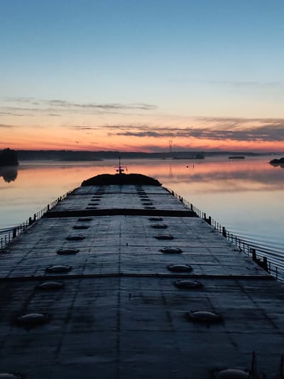 The image shows a long, flat barge on a calm body of water, possibly a river or lake, at dawn or dusk. The barge is dark, with a grid-like pattern and round hatches. Its railings are visible along the sides. The water is reflecting the sky, which is a gradient of blue at the top, fading to orange and pink near the horizon. There is a thin layer of fog or mist above the water. In the distance, there are silhouetted trees and possibly a power line. The location cannot be confirmed.