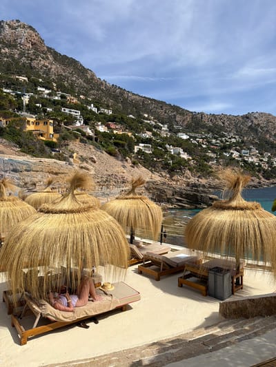 The image depicts a sun-drenched coastal scene in Port D'andratx, Spain, characterized by its hilly terrain and azure waters. In the foreground, several large, thatched umbrellas provide shade over empty lounge chairs, with one occupied by a woman in a white bikini reclining and looking at her phone. A straw hat rests on the lounge chair next to her. The beach area is paved with light-colored stone, with steps leading down to a rocky shoreline where a few people are visible. Behind the beach, the steep, verdant hillside is dotted with white and yellow buildings, characteristic of Mediterranean architecture. The sea is a vibrant turquoise, meeting the rugged coastline. The sky is partly cloudy, with fluffy white clouds against a bright blue backdrop.