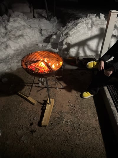 An outdoor nighttime scene in Ветеран Снт, Russia, depicts a circular, metal charcoal grill containing burning wood and glowing embers, casting an orange light. The grill stands on thin metal legs, which are stabilized by two wooden planks forming a cross-base on the gravel ground. Behind the grill and to the left, large irregular piles of snow are visible, indicating cold weather conditions. To the right of the grill, a person is partially visible, seated. This individual is wearing dark pants and distinctive bright yellow, puffy slippers. Their hands are clasped or resting on their lap. Adjacent to the person, on a darker surface, are several indistinct objects, including what appears to be a metallic can and a flat board. A white vertical post of a structure is visible on the far right. Shadows are cast by the light emanating from the grill fire.