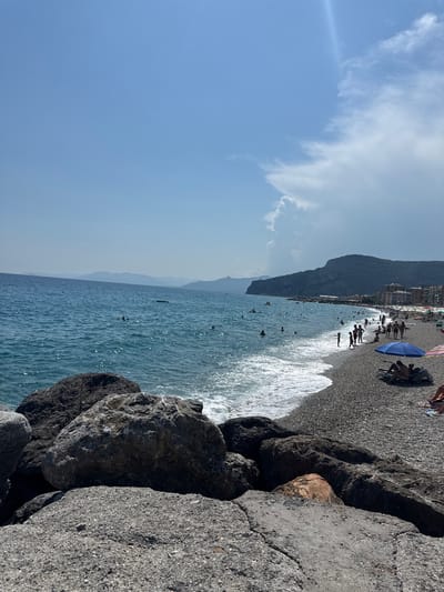 This image captures a sunny day at a beach, likely in Finale Ligure, Italy, given the characteristic pebble beach and the mountainous coastline. In the foreground, large, weathered rocks line the edge of the pebble beach, with the turquoise water of the Ligurian Sea gently lapping at the shore. The sea is moderately populated with swimmers and a few kayakers visible in the distance.

Further along the coastline, the beach is lined with rows of colorful umbrellas and lounge chairs, indicating a popular tourist destination. Numerous people are present, some sunbathing, others wading in the water, and a few children playing near the shore. Buildings, possibly hotels or apartments, are visible nestled against the hillside in the background, which is characterized by lush green vegetation and rocky outcrops. The sky is a clear, bright blue with a few wispy clouds, suggesting fair weather. The overall scene depicts a typical summer day at an Italian beach resort.