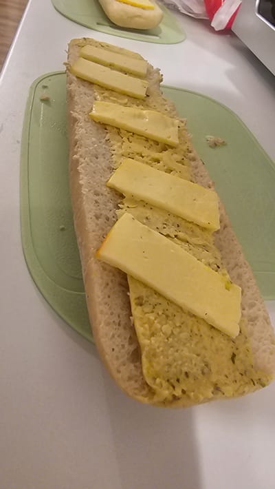 A half-eaten cheese sandwich rests on a light green cutting board on a white countertop. The sandwich, composed of what appears to be a long roll, is split open, revealing a spread of yellow paste-like substance covered with thin rectangular slices of yellow cheese. Another small bun with a similar yellow filling is visible on the left side of the image, situated on another light green cutting board. To the left of the small bun is a red and white wrapper. No people are visible in the image. The scene appears to be indoors, likely a kitchen. There is no indication of the city of Rüsselsheim am Main.