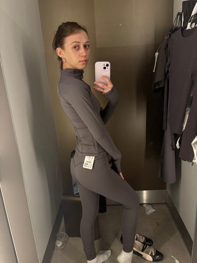 The image shows a young woman in a gray athletic outfit standing in a dressing room, taking a selfie in a mirror. She has her hair pulled back and is wearing a ring on her left hand and earrings. Her outfit consists of a long-sleeved zip-up top with thumbholes and matching leggings. A price tag is attached to her leggings. In the background, clothing items are hanging on a rack. On the floor, there are a pair of black and cream Puma ballet flats, white socks, a plastic cup, and some crumpled white paper. The dressing room appears to be in a retail store. There is no information in the image that suggests it is located in Aschaffenburg, Germany.