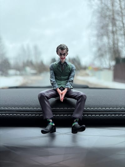 A highly detailed Joker figurine, modeled after Heath Ledger's portrayal, sits centrally on a black automotive dashboard with white stitching. The figurine wears a green vest, light blue patterned shirt, dark tie, dark purple trousers, black shoes, and colorful patterned socks, with hands clasped in front and legs spread. The background, viewed through the vehicle's windshield, depicts a blurry, overcast winter or early spring scene in Lappelovo, Russia. A muted grey sky dominates the upper frame, with a narrow asphalt road receding into the distance, flanked by bare deciduous trees and faint outlines of residential structures or fences. Patches of white, consistent with snow or slush, are visible along the road's edges.