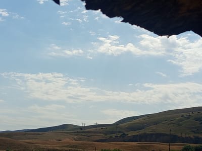 The image depicts a vast, arid landscape under a cloudy sky, with rolling hills and sparse vegetation. In the foreground, the earth appears dry and brown, possibly indicating a rural or steppe environment. The hills in the midground are covered in patches of green and brown, suggesting a mix of grasses and dry soil. Several power pylons are visible scattered across the landscape, hinting at the presence of infrastructure. In the upper portion of the image, a dark, textured object with a jagged edge obscures a small part of the sky, which could be a natural formation or part of a man-made structure. The overall impression is one of a remote, undeveloped area. The location is identified as Jardy-Su, Kyrgyzstan, a region known for its natural beauty and undeveloped landscapes.