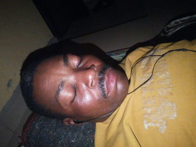 A close-up, angled shot shows a person lying down, sleeping. The person has dark skin, a mustache, and is wearing a yellow shirt. Earbuds are visible in the person's ear and draped over the shirt. The background is dark and out of focus, with a hint of a textured surface that may be a carpet or rug. The scene appears to be indoors. The image was taken in Ajasse Ipo, Nigeria.