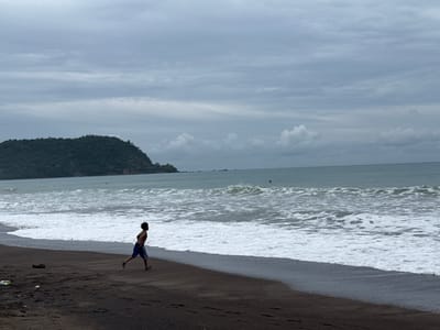 The image shows a person jogging on a dark sand beach in Jacó, Costa Rica, with the Pacific Ocean and a lush green headland in the background. The sky is overcast with light gray clouds. The person, appearing to be a male with dark curly hair and wearing blue and white striped shorts, is in mid-stride, facing left. Waves are breaking on the shore, creating a foamy white edge along the dark wet sand. A dark object, possibly a shoe, lies on the dry sand to the left of the jogger, along with some litter. The headland on the horizon is covered in dense green vegetation and slopes down to the sea. The water appears choppy with whitecaps. There are at least two small red buoys visible in the water. The overall atmosphere is moody due to the cloudy sky.