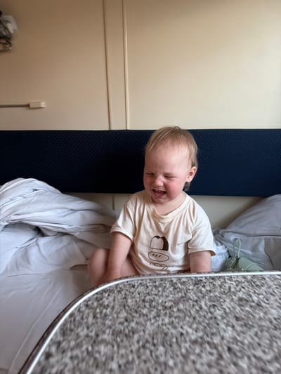 A young, light-skinned child with blonde hair, wearing a beige t-shirt with a cartoon print, is lying on a bed with white sheets and a blue cushion. The child is smiling with eyes closed. A speckled table is positioned on the right side. The setting appears to be a train compartment, evidenced by the narrow bed, the blue cushion, and the wall design. A power outlet is visible on the wall. The city of Kizner, Russia cannot be confirmed from the given image.