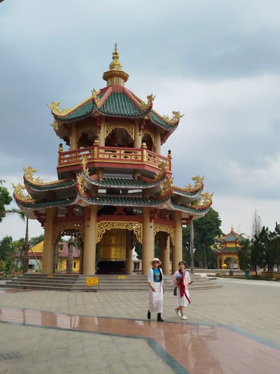 Two women are walking in front of a large, ornate, pagoda-like structure in Phú An, Vietnam. The structure has multiple tiers with green roofs and gold detailing. A large golden bell hangs in the center. One woman wears a white skirt and jacket, a blue t-shirt, a hat, and boots. The other woman wears a white skirt and sneakers with a pink jacket draped over her shoulders. The plaza is paved with patterned tiles. Another, smaller pagoda is visible in the background. The sky is overcast.