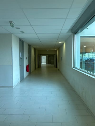 This image displays an empty, straight interior hallway within a modern institutional or commercial building, specified to be located in Montigny-lès-Metz, France.
The perspective is eye-level, looking longitudinally down the corridor. The ceiling is a grid of white suspended acoustic panels, uniformly illuminated by multiple recessed circular light fixtures. A green-and-white emergency exit sign, depicting a running figure and an arrow, is mounted on the ceiling in the upper left foreground.
The left wall, painted a light, neutral color, features several grey, solid doors spaced along its length. A red, wall-mounted fire extinguisher is prominently displayed on this wall between two doors in the mid-ground. The right wall, also painted a light, neutral color, is dominated by a large, rectangular window with a white frame. Above the window is a white housing for a roller shutter. The window reflects interior light and offers a partial, indistinct view of an exterior environment, possibly another building or a parking area.
The floor is covered with light-toned, rectangular tiles arranged in a staggered pattern, extending without interruption into the background. The corridor gradually narrows in the distance, revealing additional doorways, some partially ajar, at its far end. The overall lighting is even, casting a slightly cool tone over the scene. No individuals, personal belongings, or any ongoing activities are present, indicating the hallway is currently unoccupied.