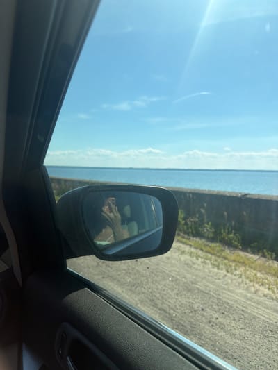 The image captures a view from inside a vehicle, looking out the passenger side window towards a body of water. In the foreground, the dark interior of the car, including the window frame and part of the door panel, is visible. A side-view mirror reflects the image of a person holding a phone, taking a selfie. Beyond the car, a calm, blue body of water stretches towards the horizon, meeting a clear, bright sky with scattered clouds. A low, stone wall runs along the edge of the water, separating it from a sandy or gravelly roadside with sparse vegetation. The scene appears to be located in Lyutizh, Ukraine, likely along the Kyiv Reservoir, given the landscape.