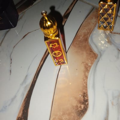 The scene depicts three containers resting on a tiled surface in Dan Maliki, Nigeria. A primary red, rectangular bottle with gold trim and an ornate gold cap, topped with a small clear gem, is centrally positioned. Its visible side features a design with red hearts and a red stylized face or emoji on a gold panel. To the right, a second rectangular bottle is partially visible, displaying a pattern of gold grids over clear or silver sections. A dark-colored object, likely another bottle, is partially visible in the upper left corner. The objects are situated on a light-colored tiled surface, exhibiting a pattern of white and grey marbling alongside a tan section with darker brown veins. A shadow is cast by the gold-capped bottle onto the tile. No individuals, actions, or interactions are present in the image.