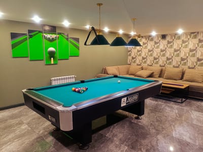 The image depicts an indoor pool hall with a green billiard table in the foreground. The table is set up with a rack of multi-colored billiard balls, and a white cue ball is also visible on the felt. Behind the pool table, a sectional couch with light brown cushions fills the mid-ground. To the left of the couch, a radiator is mounted on a pale green wall. Above the couch, a wall features a repeating geometric pattern in muted tones of beige, green, and gray. A set of three pendant lights with dark green shades hang from the ceiling over the billiard table, casting light on the scene. To the left of the pool table, a five-panel wall art displays a stylized image of a billiard table, including a cue ball and racked balls. The floor is covered in large, gray marble-patterned tiles. The overall environment suggests a recreational space. The location is identified as Kok Jar, Kyrgyzstan. There are no people visible in the image.