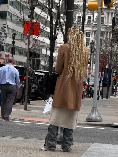 The image captures a street scene in Philadelphia, Pennsylvania, United States. In the foreground, a woman with long blonde braided hair, wearing a brown coat, a beige skirt, and dark pants, stands on the sidewalk. A man in a blue shirt and gray pants is seen walking nearby.

Behind them is a street with parked black cars and visible pedestrian crosswalk markings. A traffic light pole stands tall on the sidewalk edge. Various signs are affixed to the pole. The "DE TOUR" sign, oriented to the left, is observed. A sign warning that a vehicles yield to pedestrians is partially visible.

The background is characterized by urban architecture, including tall buildings with windows and bare trees. The sky is overcast, casting a muted light on the entire scene.