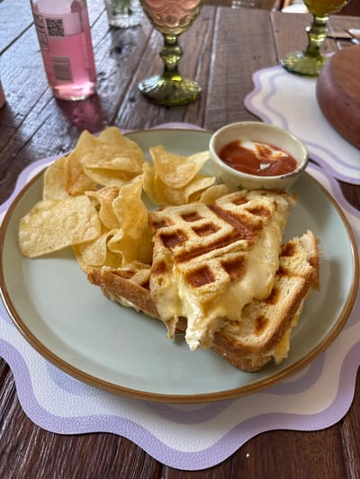 This is a top-down, close-up shot of a waffle sandwich with cheese and a side of potato chips, served on a light blue plate with a brown rim. The sandwich appears to be made with thick slices of bread pressed into a waffle iron, resulting in a golden-brown, textured exterior and melted, gooey cheese oozing out from within. A small ramekin of what appears to be ketchup or a similar red sauce with a dollop of cream or mayonnaise sits to the left of the sandwich. A pile of thinly sliced, golden potato chips is arranged around the sandwich. The plate is placed on a dark wooden table, partially covered by a decorative placemat with a wavy pattern in shades of light purple and white. In the background, out of focus, are two decorative gold-colored stemmed glasses and a small pink bottle, possibly of wine or sparkling cider. A wooden bowl is also visible in the upper left corner. The overall scene suggests a casual meal or snack, possibly at a home or a relaxed eatery. The lighting appears to be natural, suggesting it could be daytime. There is no visible text on the sandwich or chips, but the pink bottle has some indistinct text and a QR code visible on its label. The location context is Rionegro, Colombia.
