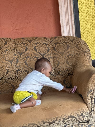 A baby in a white long-sleeve shirt and yellow shorts lies on a brown patterned couch, holding a pink object. The baby's left arm is extended, reaching towards the top of the couch. The background includes a peach-colored wall, a white ledge, and a yellow surface with a diamond pattern. The couch has an ornate floral design, with the baby positioned in the center.