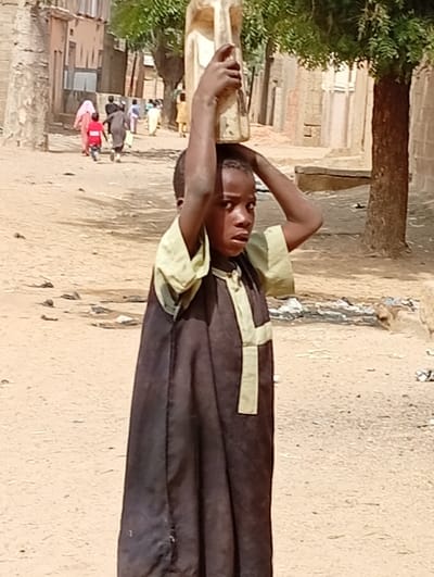 A child in a traditional brown and light green robe carries a large, light-colored plastic container on their head. They are standing on a dirt road in what appears to be a village. In the background, several other people are walking down the road, and there are buildings and trees lining the street. The lighting suggests it is a sunny day. The child has a concerned or serious expression.