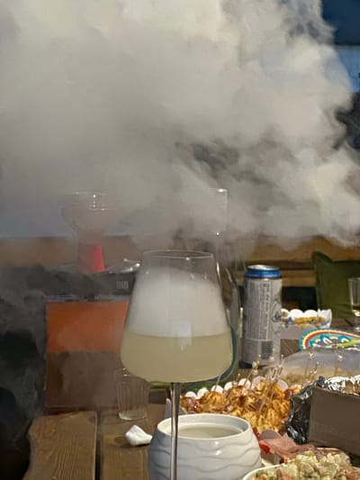 This image captures a gathering at dusk, possibly in the Тумілавіцкі сельскі Савет, Belarus region, characterized by an abundance of white smoke or vapor. A large wine glass filled with a pale yellow liquid topped with a thick layer of foam is prominently featured in the foreground, partially obscured by the swirling smoke. In the background, a table is laden with various food items, including what appears to be a platter of appetizers on skewers and a container of salad. A can of a beverage, possibly beer, is also visible on the table, alongside other glasses and what might be a hookah apparatus, which could be the source of the smoke. The overall ambiance suggests a casual social event, with the dense smoke creating a dramatic and somewhat mysterious atmosphere.
