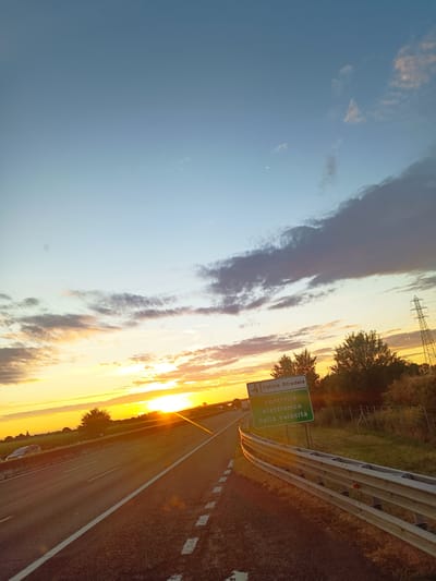 The image shows a highway at sunset. A sign reads "Polizia Stradale controllo elettronico della velocitá," which translates to "State Police electronic speed control." The sun is setting on the left side of the image, casting a warm glow over the scene. The sky is a mix of blue and orange, with some clouds. There are trees and fields on the right side of the highway. The image was taken in the city of Somaglia, Italy.