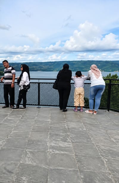 Five people stand on a tiled balcony overlooking a body of water and a forested landscape under a cloudy sky. On the left, a man wearing a black and white striped shirt and black pants leans on a railing. Next to him, a woman in a black hijab and white top, also in black pants, stands with a small bag over her shoulder. To their right, a woman in a black hijab and black pants stands facing away from the camera. In the center, a young child wearing a white shirt and light yellow pants stands between two women. The woman on the right is wearing a light pink hijab, a white shirt, and blue jeans. The background shows a wide expanse of water, bordered by green hills and a cloudy sky.
