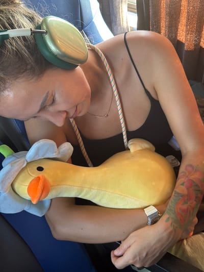 A young woman with blonde hair, wearing green headphones and a black tank top, sleeps slumped over a yellow stuffed duck. She has a colorful beaded lanyard around her neck and a watch on her left wrist. A floral tattoo is visible on her right arm. The background appears to be the interior of a vehicle, possibly a bus, with blue seats and brown curtains. The image does not provide any specific location in Hoa Xuan, Vietnam.