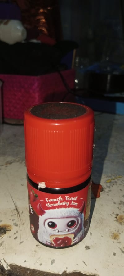 A small container with a red lid and label reads "French Toast Strawberry Jam." The label features a cartoon monster-like character with large red eyes holding a piece of toast covered in strawberry jam. The lid is cylindrical and textured. The container sits on a dirty surface, with blurred objects in the background including a pink textured item.