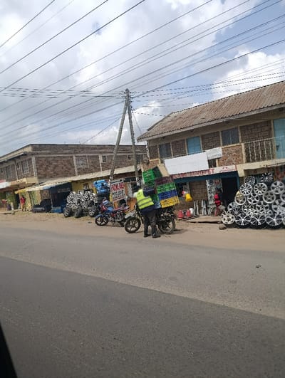 The scene depicts a commercial street in Matungulu west ward, Kenya. An asphalt road occupies the lower and middle sections of the image. The sky above is partially cloudy with patches of blue, traversed by multiple overhead power lines supported by a wooden utility pole.
On the right side of the road, a two-story building with a stone-and-plaster facade and a corrugated metal roof is visible. Its ground floor hosts a business identified by "MILAND AGROPET" signage, displaying numerous silver automotive wheel rims stacked externally. Several red plastic containers are positioned near the building's entrance, alongside additional green and yellow plastic crates.
In the mid-ground, a person wearing a yellow-green high-visibility vest over dark clothing stands next to a dark-colored motorcycle. This motorcycle is heavily loaded with multiple green and yellow plastic crates secured to its rear. A second motorcycle, red and black, is parked immediately to the left of the loaded motorcycle.
Further left along the street, another two-story building of similar construction is present, with some commercial signage visible, including partial text "DA OLD CHICKS." Various items, potentially automotive parts, are displayed outside this structure. Faintly visible individuals are present near the buildings in the background. The ground beside the road is a mix of dirt and gravel.