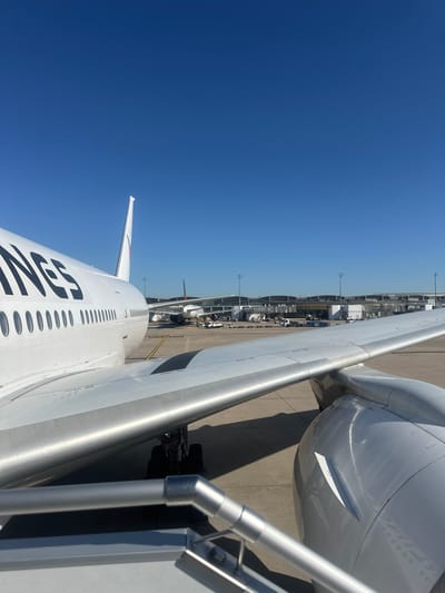 This image depicts an airplane at Charles de Gaulle Airport, located in Tremblay-en-France, France. The viewpoint is from a passenger boarding bridge, looking onto the wing and engine of the aircraft. Visible markings on the fuselage read "NAS". Another aircraft is visible further down the tarmac alongside airport terminal infrastructure. The sky is a clear blue.