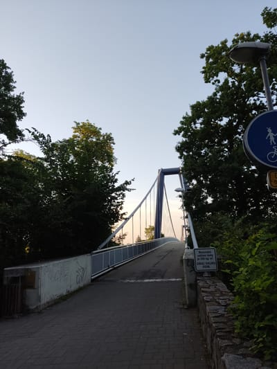 The image shows a pedestrian bridge in Kolín, Czechia. The bridge is made of steel and concrete and has a modern design with suspension cables. The walkway is paved with cobblestones. On the right side of the walkway, there is a sign with text in Czech that indicates a maximum load capacity and prohibits cycling. There are trees and greenery on both sides of the bridge. The sky is clear and the lighting suggests it is either early morning or late afternoon.