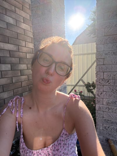 A woman with light skin and reddish-blonde hair wears glasses and a pink floral sundress. She puckers her lips for a selfie. The background shows a brick wall, a light-colored fence, and sunlight filtering through trees. The scene appears to be outdoors, possibly in a yard.