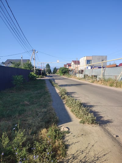 A street scene in the city of Zhaŋa Qūat, Kazakhstan, under a clear blue sky. The image is taken from a low angle, looking down a paved street lined with a sidewalk and grassy areas. On the left, a dark corrugated metal fence encloses a property with a house visible behind it. Trees and shrubs grow along the fence. Overhead, power lines stretch across the sky, supported by wooden poles. On the right side of the street, more fencing, some made of corrugated metal and supported by wooden posts, delineates properties with houses visible beyond. In the distance, the street curves slightly and leads towards a mountainous landscape. A traffic sign indicating a height limit of 4.5 meters is visible over the road. The ground on the right side of the street appears to be unpaved and sandy, with sparse vegetation. The overall impression is of a residential street in a developing area.