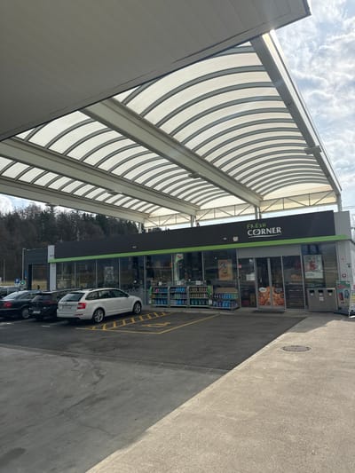 The image depicts the exterior of a "Fresh Corner" convenience store, likely part of a gas station, located in Radovljica, Slovenia. The scene is observed during daytime under an overcast sky with visible sunlight.

The primary subject is a single-story building featuring a dark gray facade with a horizontal green stripe positioned above the windows and entrance. The "Fresh Corner" logo, rendered in white text with a green leaf icon, is centered above the main entrance. The building's front is primarily composed of large glass windows showcasing various retail products and promotional signage within. A double-door entrance is centrally located.

A large, modern canopy with translucent, arched panels supported by a light-colored metal framework extends over the front of the building and an adjacent parking area. In front of the store, an asphalt parking lot contains three parked passenger vehicles: a partially obscured dark sedan on the far left, a dark hatchback/sedan, and a white Škoda Octavia Combi station wagon. The white station wagon is positioned within an accessible parking space, denoted by yellow markings and a discernible wheelchair symbol on the asphalt.

To the immediate left of the store's entrance, two metal display racks hold various bottled beverages or similar packaged goods. To the right of the entrance, two gray waste receptacles are present, alongside a small A-frame promotional sign. Security cameras are affixed to the building's facade.

In the background, beyond the building and parking area, a densely forested hillside or mountain range is visible, primarily consisting of bare trees, suggesting a non-summer season. A solitary human figure is faintly discernible walking near the left side of the building in the distance, partially obscured by the parked vehicles. The foreground consists of a concrete slab.
