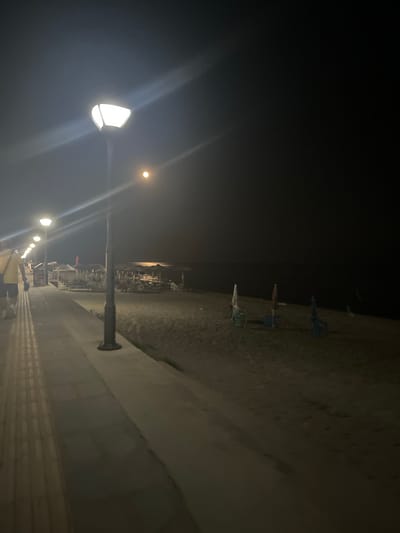 The image depicts a nighttime scene on a beach in Ορφάνι, Greece. A paved walkway, lined with tactile paving, runs along the left side of the frame. Several streetlights illuminate the walkway and the adjacent sandy beach. The beach is sparsely populated with closed beach umbrellas and chairs. The sea is visible in the background, appearing dark and calm. A person wearing a yellow shirt and dark shorts is walking away from the camera on the walkway. The overall atmosphere is serene and quiet.