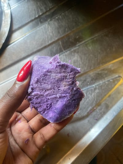 In Kianyaga, Kenya, a dark-skinned left hand with a long, pointed, bright red polished fingernail on the thumb holds a irregularly shaped piece of purple root vegetable, consistent with a cooked purple sweet potato or yam. The food item displays a fibrous, textured interior with varying shades of purple and some lighter streaks on its broken surface. The background consists of a reflective, ribbed metallic surface, likely a stainless steel kitchen sink or drainer.
