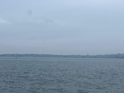 The image presents a panoramic view of Bangor, United Kingdom, observed from across a body of water. The upper portion consists of an overcast, light grey sky, featuring a dark, amorphous smudge artifact in the upper-center. The lower half is comprised of dark grey-blue water, exhibiting widespread surface ripples. Along the distant horizon, the cityscape of Bangor, UK, is visible as a continuous band of buildings and structures. Two distinct spires, characteristic of churches or tall buildings, rise above the general skyline, one near the center-right and another further to the right. A single, small, light-colored vessel, appearing as a silhouette with a visible mast or cabin, is situated on the water in the middle-ground, positioned slightly left of the image's vertical center. No human figures or specific dynamic actions are identifiable. The entire scene is affected by atmospheric haze, contributing to the subdued visibility of distant features.