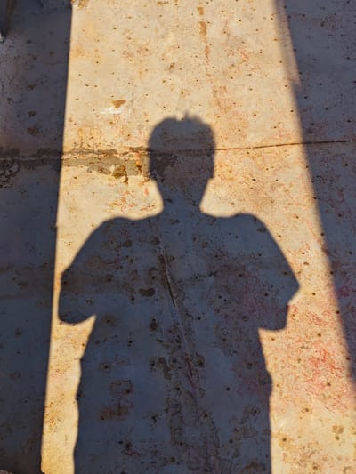 A shadow of a person with arms crossed is cast on a rusted, textured surface. The surface appears to be metal, possibly weathered or industrial, with patches of rust and peeling paint. A bright light source, likely the sun, creates a sharp, diagonal shadow that dominates the lower left portion of the image, with a second, fainter shadow to the right. The environment is outdoors, in an urban setting, with the distinct texture of the ground suggesting it might be a weathered metal platform or walkway.