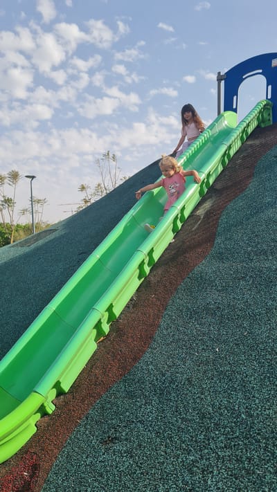Two females are on a green, covered slide at a playground. One female, presumably a child, is actively sliding down, while the other, presumably an older child or adult, is seated near the top of the slide, seemingly preparing to slide down as well. The playground is constructed on a green and brown textured surface with the slide being the central object. The image appears to be taken outdoors on a day with a partly cloudy sky. In the background, trees, a crane and street lamps are visible. The city is Kiryat Gat, Israel.