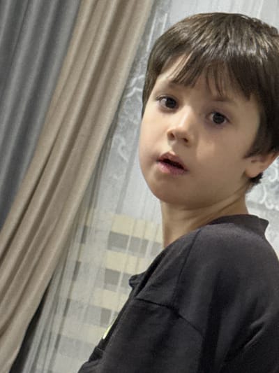 The media file is a close-up, tilted shot of a young boy. He has dark hair and appears to be looking directly at the camera with an open mouth. His left eye is more visible than his right, and his skin tone is fair. He is wearing a dark, long-sleeved shirt. The background is blurred, but appears to be an indoor setting with curtains or drapes in muted tones of beige and gray. The lighting suggests it is daytime. The image is slightly grainy, indicative of a photograph taken with a mobile device or in low light conditions. There is no visible text in the image. The overall impression is that of a candid or informal portrait.